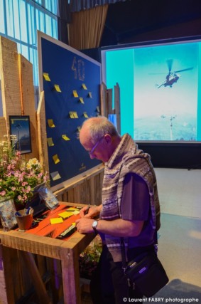photographe événementiel à Albertville : un invité laisse un petit mot sur un post-it en guise de livre d'or, alors qu'est projeté sur un écran une intervention en montagne par hélicoptère